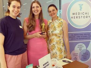 Demo Night 2023 third place winners, Medical Herstory stood next to their stand holding a trophy