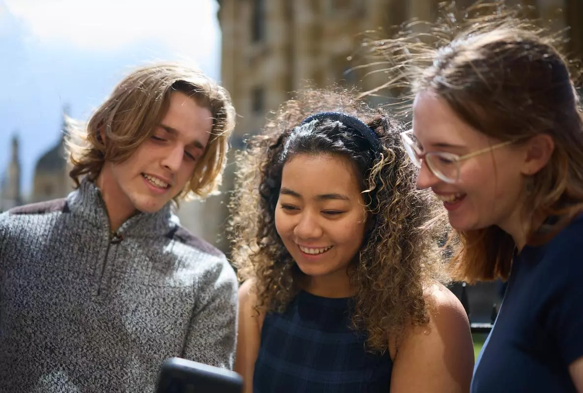 STUDENTS LOOKING AT A PHONE TOGETHER OUTSIDE THE RADCLIFFE CAMERA